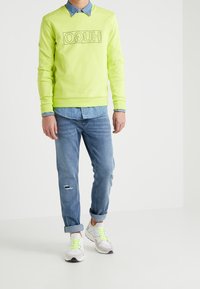 Felpa gialla neon con logo "HUGO", indossata sopra una camicia di jeans azzurri. Abbinata a jeans blu con effetti consumati e sneakers bianche con accenti neon.