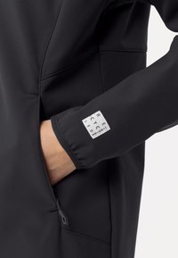 Zwarte softshell jack met een ritszak en een label met de tekst "GERECYCLEERD." De stof is glad met een matte afwerking.
