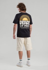 T-shirt nera in cotone con una grafica del sole e testo sul retro. Abbinata a pantaloni corti beige e scarpe bianche con calze a strisce verdi.