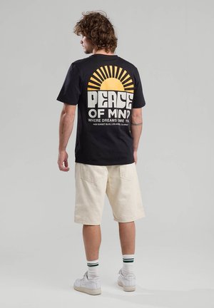 Black cotton t-shirt featuring a sun graphic with text on the back. Paired with beige shorts and white shoes with green-striped socks.