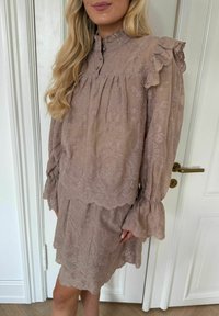 Femeie îmbrăcată într-o rochie mov pal cu dantelă, guler înalt cu volane, mâneci lungi bufante și tiv zimțat, stând lângă o ușă albă cu panouri.