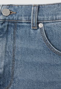 Lähikuva sinisistä farkuista, joissa näkyvät vyötärö, vyönlenkki, metallinen nappi ja osittainen etutasku näkyvine ompeluyksityiskohtineen.