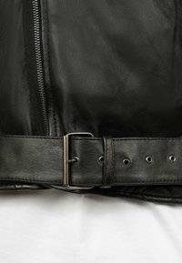 Sezione di giacca in pelle nera con una cerniera argentata e una cintura regolabile con fibbia metallica e numerosi fori.