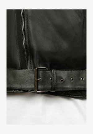 Détail d'une veste en cuir noir présentant une fermeture éclair en métal, une boucle avec une sangle et une finition texturée sur le cuir.