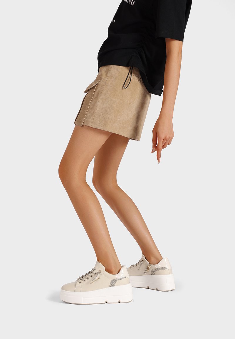 Sneaker in suede beige con dettagli testurizzati, suola platform bianca e lacci abbinati. Indossati con un top nero e una gonna beige.