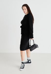 Vestido negro de punto con un detalle de lazo en la cintura, combinado con zapatillas negras y calcetines grises. Los accesorios incluyen un pequeño bolso negro.