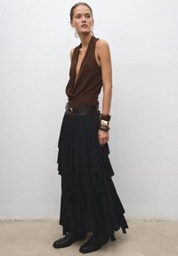 Haut marron sans manches avec un décolleté en V profond, associé à une jupe maxi noire à volants. Une ceinture noire souligne la taille ; le mannequin porte des bottines noires et des bracelets.