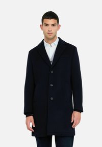 Man in een marineblauwe knopenjas over een lichtblauw shirt en grijze vest, staand tegen een effen witte achtergrond.