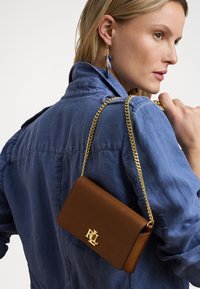 Geantă de mână texturată maro cu curea din lanț auriu și accent cu logo, purtată pe umărul îmbrăcat în blugi, evidențiind forma sa rectangulară elegantă.