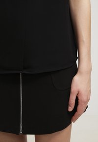 Top nero con una texture liscia, abbinato a una gonna nera con una cerniera metallica visibile frontale e dettagli sulle tasche laterali.
