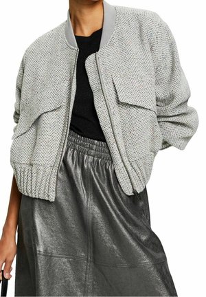 Femme portant une veste zippée texturée gris clair sur un haut noir et une jupe brillante gris foncé, tenant un sac à main noir.