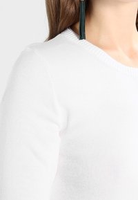 Bílý svetr s žebrovaným výstřihem a texturovanou látkou. Detailní záběr na oblast ramene a krku.