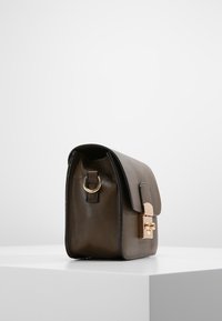 Brun skuldertaske i læder med klap, guldfarvede beslag og en metallring i siden. Glat tekstur uden synlige mønstre.