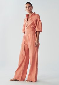 Mujer vistiendo un mono de pierna ancha coral rosa con cinturón y mangas abullonadas, de pie descalza frente a un fondo gris claro liso.