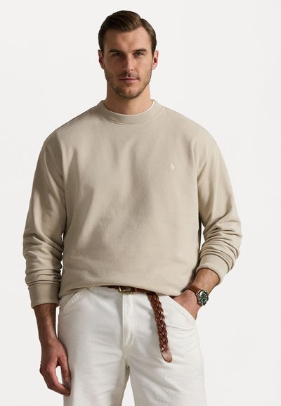 Beige sweatshirt med rund hals og ribbede mansjetter, med en liten brodert logo på brystet, kombinert med hvite bukser og et flettet belte.