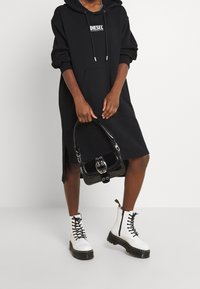 Schwarzes Kapuzenkleid mit Taschen vorne, weißem Logo und seitlichen Schlitzen; kombiniert mit weißen Schnürstiefeln und einer schwarzen Handtasche.