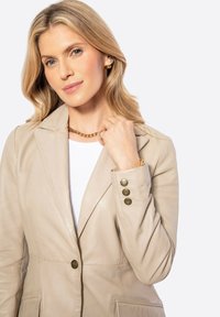 Beige leren blazer met gouden knopen en details van een manchetknoop met drie knopen; gedragen over een witte top; gecombineerd met een gouden ketting.