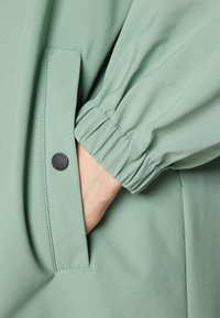 Veste vert clair avec des poignets de manches froncés et une poche latérale dotée d'une couture contrastante. Tissu doux avec une texture lisse.