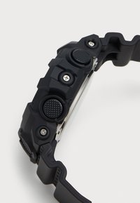 Montre en caoutchouc noir avec un boîtier texturé, trois boutons-poussoirs et des vis en acier inoxydable. Le design présente des formes audacieuses et angulaires ainsi qu'un bracelet épais.