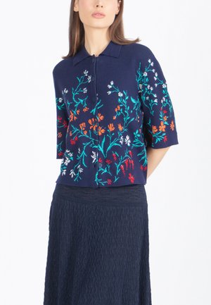 Naine, kes kannab tumesinist lühikeste varrukatega pluusi mitmevärvilise lillemustriga tikandiga ja tekstureeritud tumedat seelikut, poseerib ühevärvilise tausta ees.