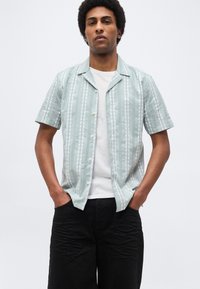 Άνδρας με τα χέρια στις τσέπες, φοράει ένα ελαφρώς πράσινο πουκάμισο με μοτίβο και κοντά μανίκια πάνω από λευκό T-shirt και μαύρο παντελόνι.