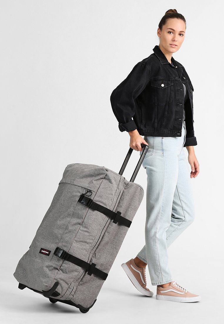 Grå stof rejsetaske med sorte stropper, lynlås og en teleskopisk håndtag, der bliver trukket af en person i lyse jeans og en sort jakke.