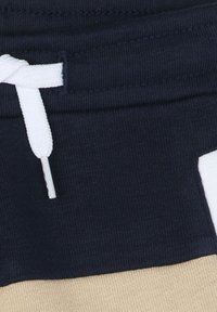 Pantaloni jogger blu navy con cordino bianco, caratterizzati da un design a blocchi di colore con accenti beige sul fondo. Tessuto morbido e testurizzato.