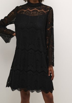 Robe longue en dentelle noire à manches longues, col montant, motifs floraux et géométriques complexes, et ourlet festonné portée par une personne debout.