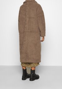 Długi brązowy płaszcz z futra syntetycznego z wysokim kołnierzem, o luźnym kroju i teksturowanej powierzchni; noszony na wzorzystej zielonej spódnicy i czarnych botkach bojowych.