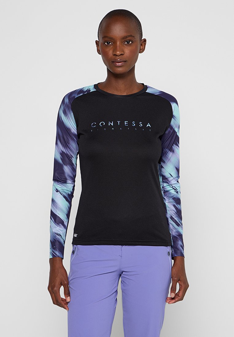 Frau in einem schwarzen Langarmshirt mit blauen gemusterten Ärmeln und dem Text "CONTESSA SIGNATURE", kombiniert mit helllila Hosen, stehend.
