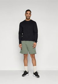 Svart genser med rund hals, grønne shorts, hvite treningssokker og svarte joggesko. Modellen står foran en nøytral bakgrunn.
