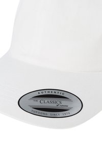 Hvid kasket med en buet skygge, udstyret med et tekstureret stof og et sort ovalt mærke, der står "Authentic THE CLASSICS YUPOONG SINCE 1974."