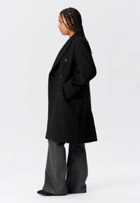Woman with braided hair wearing a black double-breasted coat and dark flared jeans, standing in profile against a plain background.