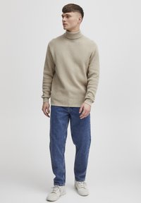 Beige textured turtleneck sweater with ribbed cuffs and hem, paired with blue denim jeans and white sneakers. Standing pose against a plain background.