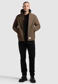 Brown, padded jacket with a high collar; black zipper and pockets; worn over a black sweater and paired with black pants and shoes.