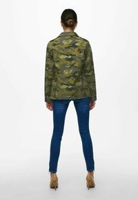 Kamuflagejakke i grønne og brune nuancer, med krave, knaplukning og justerbar talje, parret med blå jeans.