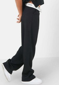 Zwarte wijde broek met een textuurafwerking, voorzien van zijzakken en een witte tailleband met het label "SIXTH JUNE." Gepaard met witte sneakers.