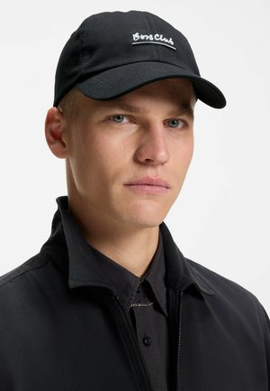 Young man with light skin and blue eyes wearing a black baseball cap with "Boss Club" text and a black collared jacket over a buttoned shirt.
