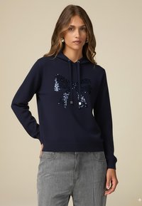 Marinblå huvtröja med en dekorativ paljettrosett i nyanser av blått. Tillverkad av texturerat tyg med ribbade muddar och nederkant.