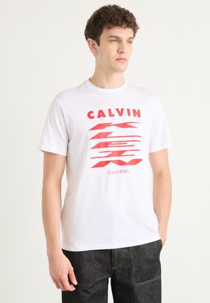 Balti medvilninė marškinė su raudonu piešiniu, ant kurio yra žodžiai "CALVIN" ir banguotos linijos. Apvalus kaklo iškirpimas ir trumpomis rankovėmis. Dėvimi tamsūs, tekstūruoti džinsai.