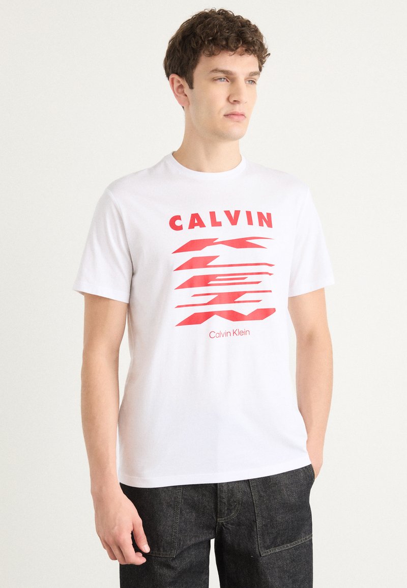 Biely bavlnený tričko s červeným grafickým potlačou s nápisom "CALVIN" a vlnitými líniami. Okrúhly výstrih a krátke rukávy. Nosia sa tmavé, textúrované džínsy.