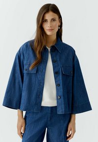 Jeansjack met een recht model, knoopsluiting, borstzak en ruime mouwen. Donkerblauwe kleur en gemêleerde stof. Draag het over een wit topje.