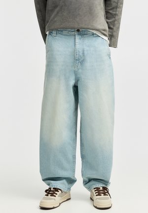 Loose light blue faded jeans worn with beige sneakers with dark laces, paired with part of a gray long-sleeve top visible above waistband.