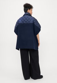 Gilet matelassé bleu marine avec un bas en tissu tricot contrastant, des manches courtes et une coupe décontractée. Il dispose de fentes latérales pour plus de liberté de mouvement.