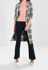 Długi, w kratkę szary i czarny płaszcz na różowej bluzce, w połączeniu z granatowymi spodniami z szeroką nogawką. Białe buty na wysokim obcasie dopełniają stylizację.