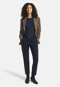Blazer met een bruin geruit patroon, marineblauw shirt en marineblauwe broek. Afgemaakt met donkerbruine enkellaarsjes en een zwarte riem. Gladde stoftextuur.