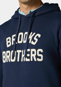 Námořnická mikina s hedvábným nápisem "BROOKS BROTHERS" v krémovém texturovaném písmu. Obsahuje kapuci se šňůrkou a bavlněnou směsovou tkaninu.