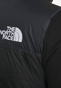 Gilet rembourré noir avec une texture lisse, arborant un logo brodé blanc sur l'épaule. Conception à col montant avec une fermeture zip.