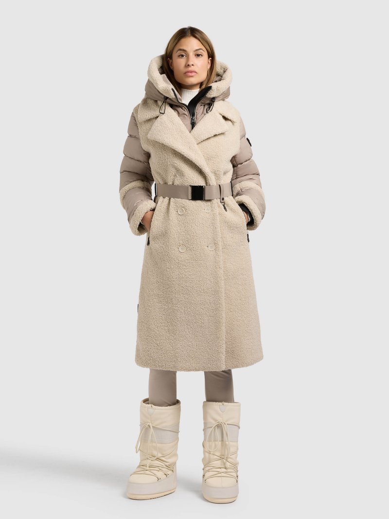 Beige dobbeltradet frakke med en blød, fluffy tekstur, stor hætte og bælte i taljen. Kombinationen omfatter beige ankelhøje støvler og tætsiddende bukser.