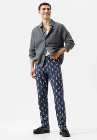 Grijze button-up shirt met een zak, gedragen over een wit T-shirt. Marineblauwe broek met kleurrijke patronen, zwarte schoenen, staande pose, neutrale achtergrond.
